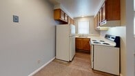 This single-floor unit offers a practical and functional layout designed to maximize comfort and usability. The living area is spacious and open, featuring neutral-toned carpet that flows seamlessly through the main rooms, creating a warm and cohesive feel throughout. Large windows fitted with horizontal blinds allow ample natural light to fill the space, enhancing the bright and airy atmosphere. The entry area transitions from hardwood-style flooring into the carpeted living space, providing a clear and welcoming threshold.

The kitchen is efficiently designed with warm honey-toned wood cabinetry both above and below the countertops, offering generous storage. White appliances, including a refrigerator and a range with cooktop, are integrated into the layout alongside a sink positioned beneath a window. Wood-look flooring in the kitchen adds durability and visual contrast to the surrounding carpeted areas.

The bathroom is clean and well-appointed, featuring a full soaking tub, a wood-vanity with a white countertop and undermount sink, a mirror, and wood-look tile flooring that complements the kitchen's aesthetic. Multiple additional rooms with plush carpet and neutral gray walls provide versatile spaces suitable for bedrooms or a home office. Throughout, the unit maintains a consistent, understated palette of soft grays and warm wood tones.