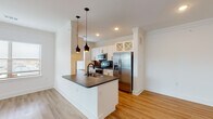 This well-appointed single-floor residence showcases a thoughtfully designed open-concept layout, where the kitchen serves as a central highlight. The kitchen features crisp white shaker cabinetry with matte black hardware, dark granite countertops, a stainless steel appliance suite including a French door refrigerator and range, a dark tile backsplash, and a peninsula with pendant lighting above — creating an inviting and functional cooking space. A hallway connects the main living areas seamlessly, with light-toned hardwood flooring running throughout, lending warmth and visual continuity across the space. Crown molding adds a refined architectural detail to the living areas. The living room is generously proportioned, offering an open canvas for flexible furnishing arrangements. Bedrooms are bright and airy, benefiting from natural light through blinds-fitted windows, ceiling fans, and clean white walls. The bathroom is a standout feature, boasting a granite vanity, matte black fixtures, and a dramatic dark marble-tiled shower and tub surround that brings bold, sophisticated contrast to the otherwise neutral palette. A distinctive dark louvered interior door adds a unique design accent throughout the space.