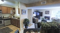 This single-floor residence offers a thoughtfully designed open-concept layout that seamlessly connects the kitchen, dining, and living areas into a cohesive and functional living space. The kitchen features rich dark wood cabinetry, a double sink, and open shelving, providing ample storage and a warm, inviting aesthetic. A half-wall partition gently separates the kitchen from the adjoining spaces while maintaining an open sightline throughout.

The dining area flows naturally from the kitchen and is furnished to accommodate comfortable everyday dining, with easy access to the living room beyond. The living room is a spacious and welcoming retreat, anchored by a classic fireplace and a built-in entertainment center with dark wood cabinetry. A textured ceiling and neutral beige walls create a cohesive backdrop throughout the shared areas.

Flooring transitions from hardwood-style in the kitchen and entry areas to plush carpeting in the living room, adding warmth and comfort underfoot. Decorative greenery and tasteful artwork punctuate the space, enhancing its livable, welcoming character. Recessed vents and ample overhead lighting ensure comfort and practicality. Overall, the space presents a balanced blend of functionality and warm, classic design.