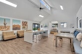Located at 650 Ebbcreek Drive in Corona, California, this single-floor community lounge and kitchen space offers a bright, open-plan layout designed for gathering and collaboration. The most striking architectural feature is the dramatic vaulted ceiling, which rises steeply above the main area, enhancing the sense of volume and airiness well beyond what the footprint might suggest. A ceiling fan and exposed beams add character to the lofty overhead space, while skylights and a generous array of windows flood the interior with natural light and offer views of the surrounding outdoor areas.

Light wood-plank flooring runs throughout, lending warmth and continuity across the space. A large central island with bar seating in teal and black serves as the focal point of the kitchen and social area, complemented by a rustic wood communal dining table with built-in banquette seating along one wall, appointed with blue and patterned cushions.

The living area features low-profile tan lounge chairs arranged beneath a triptych of bold desert landscape artwork, creating a relaxed, Southwest-inspired ambiance. One wall showcases a dynamic abstract mural in soft blues, whites, and terracotta tones. A wall-mounted flat-screen television and small-space greenery add functional and decorative touches. Black-framed glass doors open directly to the outdoors, further connecting the interior to its surroundings.