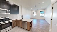 Located at 586 Central Avenue in East Orange, NJ, this thoughtfully designed single-floor unit offers a contemporary living experience with high-quality finishes throughout. The open-concept kitchen and living area serves as the heart of the space, featuring a peninsula with a sleek white quartz countertop, two-tone cabinetry in rich wood and deep charcoal tones, a full suite of stainless steel appliances including a gas range, built-in microwave, refrigerator, and dishwasher, and large-format porcelain tile flooring. The living area extends naturally from the kitchen, bathed in natural light from dual windows and complemented by recessed lighting and light-toned hardwood flooring. Additional rooms include two closets, a dedicated laundry area, and a hallway connecting the space efficiently. The bathroom is well-appointed with floor-to-ceiling white tile, a warm wood accent wall, a modern vanity with a white countertop, a soaking tub, and a large mirror with stylish wall-mounted lighting. Clean white walls, crisp trim, and recessed lighting unify the space, creating a bright and modern ambiance throughout. Urban views of the East Orange streetscape are visible through the windows.