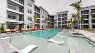 Located at 526 Vistera Blvd in Venice, Florida, this outdoor amenity space is a masterfully designed resort-style courtyard set within a contemporary multi-story residential community. The centerpiece is a generously sized swimming pool with crystal-clear turquoise water, framed by a broad, elegantly paved sun deck lined with red-and-white lounge chairs and shaded umbrella stations. Sleek in-pool loungers add a luxurious touch, allowing residents to relax directly in the water.

Surrounding the pool, the courtyard is thoughtfully landscaped with tall, swaying palm trees, lush tropical plantings, and decorative black volcanic rock beds, evoking a true Florida resort ambiance. A dedicated lawn area features a cornhole game setup and contemporary outdoor seating clusters, providing an inviting space for casual recreation and social gatherings.

An outdoor bar counter with geometric tile detailing and modern bar seating offers a stylish spot for entertaining, while the covered patio areas provide shade along the building's ground-floor corridors. The architecture of the surrounding building showcases a coastal-inspired palette of soft grey and white, with screened balconies, large windows, and arched entryways that frame the courtyard beautifully. The entire space blends functionality and aesthetic appeal, creating a vibrant, community-centered outdoor environment.