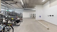 This dedicated bike storage facility is thoughtfully designed to accommodate a wide variety of cyclists and bicycle types. The space features a combination of multi-tier freestanding steel racks and wall-mounted vertical hooks, maximizing storage capacity while keeping the area orderly and accessible. The polished concrete flooring is durable and easy to maintain, well-suited for the high-traffic demands of an active cycling community. Exposed concrete ceilings with visible ductwork and conduit give the space an industrial character, while linear LED lighting runs throughout, ensuring the area is well-lit at all hours. A Luxer package storage unit is conveniently integrated into the space, adding a practical amenity for residents. Large floor-to-ceiling windows along one wall allow natural light to filter in and provide views to the outdoors, preventing the space from feeling closed off. The generous floor plan offers ample maneuvering room for bringing bikes in and out, accommodating everything from standard road bikes and cruisers to larger cargo bikes and children's bicycles. The overall layout is clean, functional, and purpose-built to support an active, bike-friendly lifestyle.