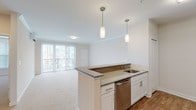 This single-floor residence offers a thoughtfully designed open-concept layout that seamlessly connects the kitchen and living area. The kitchen is a standout feature, equipped with white shaker cabinetry, granite-style countertops, stainless steel appliances including a refrigerator, range, and microwave, and a peninsula island with an integrated sink and pendant lighting overhead. Warm hardwood-style flooring anchors the kitchen and entry areas, transitioning to plush carpeting throughout the living room and bedrooms.

The spacious living area is bright and airy, benefiting from a large sliding glass door that opens to a private balcony, allowing generous natural light to fill the room. Crown molding adds a refined architectural touch. A wall-mounted split air conditioning unit provides efficient climate control.

The bedroom is generously sized with soft carpeting, a window fitted with blinds, and its own wall-mounted climate unit. The bathroom is clean and functional, featuring a full soaking tub with a shower, chrome fixtures, and wood-style flooring. A connected hallway ties the rooms together in a cohesive and practical flow, making this a comfortable and move-in-ready living space.