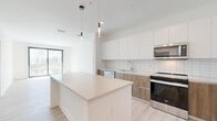 This single-floor residence offers a thoughtfully designed layout across three bedrooms and two bathrooms, with a contemporary aesthetic defined by light wood-tone flooring, crisp white walls, and clean-lined finishes throughout. The open-concept kitchen serves as a central feature, showcasing a large island with a quartz countertop, flat-front white upper cabinetry, warm wood-toned lower cabinets, a subway tile backsplash, and a full suite of stainless steel appliances including a range, microwave, dishwasher, and refrigerator. Pendant lighting above the island adds a modern accent. The kitchen flows naturally into the living area, which benefits from floor-to-ceiling vertical blinds and a sliding glass door that frames outdoor water views. The generously sized primary bedroom receives abundant natural light and connects to a well-appointed bathroom featuring a wood-vanity, white countertop, and a tiled tub-shower combination. Secondary bedrooms include built-in closets with sliding doors. A second bathroom mirrors the modern design language with vertical tile work and a soaking tub. A dedicated in-unit laundry closet houses a full-size washer and dryer, adding everyday convenience to this well-appointed home.
