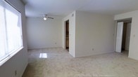 This single-floor residence offers a clean, understated aesthetic built around neutral tones and an open, functional layout. The main living area is finished with wall-to-wall carpet in a soft, warm hue that lends a comfortable and inviting feel underfoot. Smooth, light-colored walls keep the space bright and versatile, providing a blank canvas well-suited to a variety of interior styles. A ceiling fan with an integrated light fixture is centrally positioned, ensuring both air circulation and ambient lighting throughout the room. Natural light filters in, casting a soft glow across the floor and enhancing the airy quality of the space. A transitional area separates the living space from what appears to be a kitchen or utility zone, where warm-toned wood cabinetry is visible, adding a touch of classic character. The tiled flooring in this transition area contrasts neatly with the surrounding carpet, helping to define distinct functional zones. The overall layout is straightforward and efficient, with no unnecessary partitions, allowing for flexible furniture arrangements. The space presents a move-in-ready foundation that is easy to personalize and maintain.
