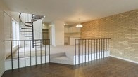 This two-story space showcases a thoughtfully designed split-level layout that creates distinct zones while maintaining an open, airy feel. The upper level features rich, dark hardwood flooring and is anchored by a sleek metal railing that overlooks the sunken lower living area, which is finished with plush carpeting for a comfortable contrast in texture.

A standout architectural detail is the full-height exposed brick wall, which adds warmth and character to the interior. The kitchen area is positioned at the far end of the lower level, equipped with white cabinetry and a peninsula that provides both workspace and casual seating. A classic chandelier hangs above, lending an elegant touch to the kitchen space.

A dramatic spiral staircase connects the two floors, serving as both a functional element and a visual focal point. The white walls throughout keep the space feeling bright and clean, complementing the varied materials and textures present in the flooring and brick accents. The combination of hardwood, carpet, brick, and metal railings gives the space a layered, well-appointed character suited to a variety of uses.