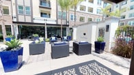 This well-appointed outdoor courtyard serves as a central amenity space within a modern multi-story residential community. The open-air patio is designed for both relaxation and social gatherings, featuring a cohesive arrangement of wicker lounge furniture with navy blue cushions, centered around a sleek glass-top fire pit table. Vibrant cobalt blue ceramic planters, filled with striking agave and tropical foliage, are strategically placed throughout the space, adding bold color and a resort-like ambiance. Tall palm trees provide natural shade and reinforce the warm, Southern California aesthetic.

The courtyard is framed by the building's contemporary façade, which combines neutral stucco tones with clean architectural lines, large windows, and private balconies above. Expansive floor-to-ceiling glass sliding doors connect the outdoor patio seamlessly to an interior clubroom, which is visible through the glazing and appears to offer additional dining and lounge seating. Decorative landscaping beds line the base of the building, enhancing the polished, manicured feel of the space. The ground is finished with large-format light stone tiles, and a decorative laser-cut patterned mat adds an artistic touch near the entrance, completing the refined and inviting character of this outdoor amenity.