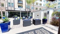 This well-appointed outdoor courtyard serves as a central amenity space within a modern multi-story residential community. The open-air patio is designed for both relaxation and social gatherings, featuring a cohesive arrangement of wicker lounge furniture with navy blue cushions, centered around a sleek glass-top fire pit table. Vibrant cobalt blue ceramic planters, filled with striking agave and tropical foliage, are strategically placed throughout the space, adding bold color and a resort-like ambiance. Tall palm trees provide natural shade and reinforce the warm, Southern California aesthetic.

The courtyard is framed by the building's contemporary façade, which combines neutral stucco tones with clean architectural lines, large windows, and private balconies above. Expansive floor-to-ceiling glass sliding doors connect the outdoor patio seamlessly to an interior clubroom, which is visible through the glazing and appears to offer additional dining and lounge seating. Decorative landscaping beds line the base of the building, enhancing the polished, manicured feel of the space. The ground is finished with large-format light stone tiles, and a decorative laser-cut patterned mat adds an artistic touch near the entrance, completing the refined and inviting character of this outdoor amenity.