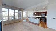 This single-floor residence showcases an open-concept layout that seamlessly connects the living area and kitchen, creating a spacious and functional environment. The kitchen is a standout feature, appointed with rich dark-stained cabinetry that contrasts elegantly against dark granite countertops. A central island with a built-in sink provides additional workspace and serves as a natural dividing point between the kitchen and living areas. Stainless steel appliances, including a range and microwave, complement the modern aesthetic, while three pendant lights hang above the island, offering both task lighting and a refined design touch. Recessed lighting is distributed throughout the ceiling, ensuring consistent illumination across the space. The living area transitions from hardwood flooring to plush carpeting, defining the two zones while maintaining visual cohesion. A bank of large windows along the far wall floods the interior with abundant natural light and provides views of the surrounding neighborhood. The overall ambiance is clean, contemporary, and well-appointed, offering a comfortable and stylish everyday living environment.