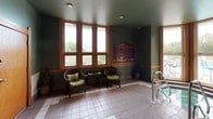 This single-floor aquatic amenity space offers a well-appointed indoor pool and hot tub area designed for comfort and functionality. The room is finished with neutral tile flooring in a grid pattern, providing a clean, slip-resistant surface throughout. Sage green walls, accented by a warm wood chair rail, create a calm and inviting atmosphere, while a deep green ceiling adds a cohesive, polished look overhead.

Large wood-trimmed windows line the walls, flooding the space with natural light and offering pleasant views of the surrounding outdoor landscape. A dedicated seating area features dark wicker chairs with green cushions and a small side table, providing a comfortable spot to relax before or after a swim. Potted plants add a touch of greenery to the environment.

The hot tub is accessible via stainless steel handrails and is conveniently positioned near the windows. An entry door with glass panels and wood framing connects the pool area to adjacent spaces. On the opposite end of the room, two wood-framed doors lead to changing or restroom facilities. Recessed lighting and posted safety signage round out the practical, well-organized design of this amenity space.