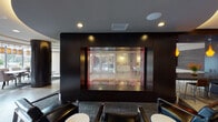 This single-floor space presents a thoughtfully designed communal lounge and lobby area with a contemporary aesthetic. The layout is organized into several distinct zones that flow seamlessly into one another, offering both open gathering areas and more intimate seating arrangements.

A welcoming entry corridor features large glass double doors with sleek metal hardware, opening into the main lounge. Adjacent to the entrance is a built-in credenza with a striking orange countertop, white cabinetry, and herringbone-patterned tile accents on the walls, illuminated by a globe pendant light. A textured three-dimensional wall panel adds further visual depth to this area.

The main lounge is anchored by a bold dark feature wall housing a large built-in display window that offers a view into an adjacent space, framed by recessed lighting. Plush seating, including a generously sized sectional sofa adorned with colorful patterned throw pillows, is arranged around a round coffee table atop a circular area rug. Curated contemporary artwork adorns the neutral-toned walls throughout, contributing to the space's gallery-like quality.

A separate dining nook with bar-height seating is visible adjacent to the lounge, accompanied by warm pendant lighting. Recessed ceiling lights and floor lamps provide layered illumination throughout, enhancing the refined, welcoming ambiance of this communal space.
