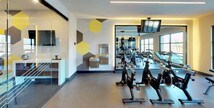 This contemporary fitness center is thoughtfully designed to support a wide range of workout routines in a single, open-plan space. The layout is anchored by a cardio zone featuring multiple treadmills and elliptical machines, all positioned to take advantage of large, floor-to-ceiling windows that flood the room with natural light and provide views of the surrounding outdoor area. A dedicated free weights section includes a dumbbell rack and an adjustable bench, complemented by a hanging heavy bag for boxing and functional training. A row of stationary cycling bikes lines one wall, reflected in a large mirror that enhances the sense of space. The walls are decorated with bold, oversized hexagonal graphic murals in yellow, gray, and charcoal, lending the space a vibrant and energetic aesthetic. Linear LED lighting runs along the ceiling, providing even, modern illumination throughout. A glass-enclosed alcove, separated by a sliding glass partition, houses a workstation with monitors — ideal for virtual training or administrative use. A built-in water fountain station adds convenience. The durable flooring transitions between rubberized and carpet-style surfaces across different zones, reinforcing the functional character of this well-appointed fitness facility.