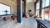 This single-floor communal space showcases an impressive industrial-modern design defined by soaring double-height ceilings and expansive floor-to-ceiling windows that flood the interior with natural light and offer views of the surrounding greenery. Exposed light brick walls serve as a prominent architectural feature throughout, contrasting warmly against dark charcoal structural columns and walls.

The main lounge area is anchored by a seating arrangement featuring patterned orange and white upholstered chairs, leather sofas, and a rustic wood coffee table, all set atop a textured area rug. A wall-mounted flat-screen television is centered on one of the brick accent walls, flanked by a wood credenza and complementary lighting. Sputnik-style chandeliers and Edison bulb wall sconces provide stylish ambient lighting throughout.

A separate seating nook features a navy leather sofa beneath a large-scale framed jet aircraft photograph, adding a bold, eclectic focal point. A generously sized communal dining area includes a heavily distressed reclaimed wood table surrounded by dark cross-back chairs, positioned beneath large windows with garden views and decorative antler wall mounts. A foosball table adds a recreational element to the space. Light gray wood-look flooring unifies the entire layout, contributing to an open, contemporary ambiance.