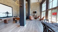 This single-floor communal space showcases an impressive industrial-modern design defined by soaring double-height ceilings and expansive floor-to-ceiling windows that flood the interior with natural light and offer views of the surrounding greenery. Exposed light brick walls serve as a prominent architectural feature throughout, contrasting warmly against dark charcoal structural columns and walls.

The main lounge area is anchored by a seating arrangement featuring patterned orange and white upholstered chairs, leather sofas, and a rustic wood coffee table, all set atop a textured area rug. A wall-mounted flat-screen television is centered on one of the brick accent walls, flanked by a wood credenza and complementary lighting. Sputnik-style chandeliers and Edison bulb wall sconces provide stylish ambient lighting throughout.

A separate seating nook features a navy leather sofa beneath a large-scale framed jet aircraft photograph, adding a bold, eclectic focal point. A generously sized communal dining area includes a heavily distressed reclaimed wood table surrounded by dark cross-back chairs, positioned beneath large windows with garden views and decorative antler wall mounts. A foosball table adds a recreational element to the space. Light gray wood-look flooring unifies the entire layout, contributing to an open, contemporary ambiance.