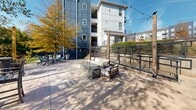 Located at 119 Pearl Ave in Greenville, South Carolina, this outdoor amenity space is a well-appointed deck and patio area designed for community gathering and relaxation. The space is divided into distinct zones that cater to a variety of social settings. A generously sized composite wood deck offers multiple high-top dining tables with chairs, ideal for casual outdoor dining or socializing, all bathed in abundant natural sunlight and framed by mature trees displaying vibrant seasonal foliage. Wooden posts throughout support overhead string lights, lending the space a warm, inviting ambiance that extends into the evening hours. Adjacent to the deck, a paved patio area features a comfortable lounge seating arrangement centered around a modern fire table, creating an intimate setting perfect for unwinding. Additional seating, including Adirondack-style chairs, is scattered throughout for flexible use. Black metal cable and wire-panel fencing defines the perimeter, providing an open feel while maintaining a sense of enclosure. Stairs provide seamless transition between levels, and the contemporary multi-story building serves as a striking architectural backdrop. The overall atmosphere is modern, relaxed, and social, making this outdoor space a standout communal amenity.