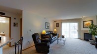 This single-floor residence offers a comfortable and functional layout designed for everyday living. The living room serves as the heart of the space, featuring plush wall-to-wall carpeting that adds warmth underfoot and a soft, inviting atmosphere. Large sliding glass doors flood the room with natural light, creating an airy, open feel while providing a direct connection to the outdoors. The neutral-toned walls offer a versatile backdrop, complemented by warm accent lighting. Framed artwork adorns the walls, adding a personal touch to the space. The room comfortably accommodates a full seating arrangement, including a reclining chair and sofa, centered around a sleek glass coffee table — ideal for relaxing or entertaining. A wall-mounted thermostat and ceiling vents indicate a climate-controlled environment for year-round comfort. The layout flows naturally, with open sightlines that enhance the sense of space. Overall, the residence presents a clean, well-maintained interior with a practical, cozy character suited for comfortable day-to-day living.