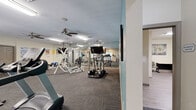 Located at 420 Riverwood Dr in Crestview, Florida, this well-appointed community facility offers a comprehensive fitness and business center experience across a single floor. The expansive main fitness area is highlighted by vibrant teal accent walls, durable carpet tile flooring, and multiple ceiling fans that keep the space comfortable during workouts. The gym is generously equipped with a multi-station cable weight machine, treadmills, ellipticals, a recumbent bike, and free weights, providing a full range of strength and cardio training options. Large windows along one wall invite natural light and views of the surrounding greenery, enhancing the workout environment. Wall-mounted mirrors amplify the sense of space throughout the fitness area. A water dispenser is conveniently located near the cardio equipment. The facility also includes a sauna, a closet for storage, and a connecting hallway. Adjacent to the fitness area, a dedicated business center features light wood-look flooring, multiple computer workstations, bookshelves stocked with reading materials, and ample seating — ideal for residents seeking a quiet, productive workspace. The overall design is clean, functional, and welcoming, making this a versatile amenity space suited to both physical wellness and everyday productivity needs.