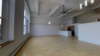 This single-floor loft-style space makes a strong first impression with its dramatic ceiling height, defined by a striking exposed beam structure that adds both architectural character and an industrial-chic aesthetic. The expansive open-plan layout provides a generous sense of space, with light hardwood flooring running throughout that enhances the room's warmth and brightness.

Large windows along one wall flood the interior with abundant natural light, complementing the clean white walls and contributing to an airy, welcoming ambiance. A ceiling fan with a brushed metal finish is centrally positioned, providing both function and a modern design accent, while a sleek pendant light fixture adds a contemporary touch overhead.

The kitchen is thoughtfully integrated into the space via a partial dividing wall that creates a natural separation without sacrificing the open feel. It features wood-toned cabinetry, stainless steel appliances including a refrigerator and microwave, and practical countertop space. A baseboard heating unit runs along the windowed wall, ensuring year-round comfort.

Overall, the space combines industrial loft character with clean, modern finishes, making it a versatile and visually compelling environment suited to a variety of uses and lifestyles.
