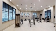 This fitness center offers a clean, modern environment designed to support a full range of workout routines. The space is finished with light-toned walls and a herringbone-patterned tile floor, creating a bright and welcoming atmosphere. Recessed lighting throughout the ceiling ensures consistent illumination across the entire floor, complemented by dual ceiling fans that promote air circulation during workouts. Multiple windows fitted with blinds line the walls, allowing natural light to filter in while offering adjustable light control. The equipment layout is thoughtfully organized, featuring a row of treadmills positioned centrally for cardio training, alongside a cable machine system from Precor for versatile strength exercises. Additional strength training machines are arranged along the perimeter, including a seated leg press and a cable-based resistance unit, with a wall-mounted television adding an entertainment option. The open floor plan provides generous spacing between machines, allowing users to move freely and safely between stations. The overall design balances functionality with a clean aesthetic, making it a practical and comfortable facility for daily fitness use.