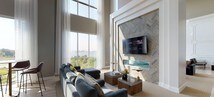 Located at Bay Drive in Bayonne, New Jersey, this thoughtfully designed single-floor space blends contemporary elegance with classic architectural details. The most commanding feature is the double-height ceiling in the main lounge area, where floor-to-ceiling windows flood the interior with natural light and frame sweeping waterfront and greenery views. Sheer drapery softens the expansive glazing while preserving the open, airy atmosphere.

The main seating area centers around a herringbone-tiled fireplace wall with a mounted television, creating a refined focal point. Light hardwood-style flooring runs throughout, lending warmth and continuity across the space. Crisp white wainscoting with geometric paneling detail adorns multiple walls, adding a tailored, sophisticated character.

A secondary lounge zone features additional seating arrangements bathed in sunlight from corner floor-to-ceiling windows offering picturesque outdoor views. A dedicated wet bar area includes a tiled backsplash, stainless steel fixtures, pendant lighting, and a generous counter with bar seating—ideal for entertaining.

Throughout, the design balances modern furnishings with classic architectural elements, resulting in a polished, welcoming environment suited for both gathering and relaxation.