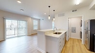 Located on Longs Peak Street in Brighton, Colorado, this one-bedroom, one-bathroom residence offers a thoughtfully designed open-concept layout with modern finishes throughout. Light wood-tone flooring flows through the main living areas, anchoring the bright and airy atmosphere created by large windows and recessed lighting.

The kitchen serves as a central hub of the space, featuring a peninsula island with a stainless steel sink, light quartz countertops, crisp white cabinetry with brushed hardware, a subway tile backsplash, and stainless steel appliances including a range and over-the-range microwave. Stylish pendant lights hang above the island, adding a refined touch. The kitchen transitions seamlessly into the living room, which is highlighted by a sliding glass door that provides direct outdoor access and fills the room with natural light.

The bedroom is a comfortable retreat with plush carpet, a ceiling fan, and an abundance of natural light from a bank of oversized windows fitted with blinds. An adjacent walk-in closet offers dedicated shelving and rod storage. The full bathroom features gray cabinetry, a wide vanity with a framed mirror, and a soaking tub. A dedicated laundry area and hallway complete the functional layout of this well-appointed Brighton residence.