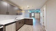 This thoughtfully designed single-floor unit blends modern finishes with industrial charm across a layout that includes a kitchen, living room, bathroom, and hallway. The open-concept kitchen is a standout, featuring dark wood cabinetry with matte black hardware, crisp white countertops, a subway tile backsplash, stainless steel appliances, and pendant lighting that adds warmth. A peninsula extends the workspace while enhancing the flow into the living area. Light wood-tone flooring runs throughout, tying the spaces together seamlessly. The living room is defined by an exposed brick accent wall with large windows that invite natural light and frame outdoor views. A striking white barn door adds character and serves as a functional architectural detail. Exposed metal ductwork along the ceiling reinforces the industrial aesthetic. The bathroom is clean and contemporary, featuring a spacious glass-enclosed walk-in shower with white subway tile surround and polished chrome fixtures. A generous open closet with built-in rod and shelving provides practical storage. The hallway connects the rooms efficiently, maintaining an open and airy feel throughout this well-appointed space.