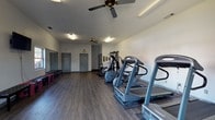 Located at 105 Lisa Street in Rincon, Georgia, this single-floor fitness center offers a well-equipped and functional workout environment designed to support a range of exercise routines. The main floor features durable wood-look flooring throughout, white walls, and a textured ceiling with ceiling fans that help maintain airflow and comfort during workouts.

The cardio section is equipped with multiple treadmills and elliptical machines, each positioned on protective rubber mats and arranged along one side of the room for efficient use of space. Alongside these, a multi-station cable and weight machine provides a comprehensive strength training option, accommodating a variety of resistance exercises in a single unit. A standalone weight bench completes the strength training area.

At one end of the facility, a open flex area features large wall-mounted mirrors ideal for form checks during stretching or bodyweight exercises. Red and black padded benches line the mirrored wall, and a wall-mounted television adds entertainment for those using the space. Natural light enters through multiple windows, offering views of the surrounding outdoor greenery.

An adjacent area serves as a wellness resource zone, featuring a standing scale, a bookshelf stocked with health and fitness reading materials, and additional seating. The space also includes interior doors leading to separate utility or storage rooms, and safety features such as an exit sign and a security camera are present throughout.