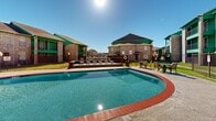 This multifamily residential complex in Houston, Texas, showcases a harmonious blend of architectural design and recreational facilities. The property features multiple three-story apartment buildings constructed with light-colored brick and distinctive green trim, creating a cohesive and visually appealing aesthetic. 

The centerpiece of the complex is a expansive, irregularly-shaped swimming pool with crystal-clear water. The pool is bordered by a red brick coping, adding a touch of classic design to the modern amenity. Surrounding the pool area, residents can enjoy various leisure facilities including comfortable lounge chairs, tables with umbrellas, and a covered cabana structure, perfect for socializing or relaxing in the shade.

The apartment buildings themselves offer private balconies, allowing residents to enjoy outdoor space from the comfort of their homes. The complex is enclosed by decorative metal fencing, enhancing security while maintaining an open feel. Well-maintained lawns and scattered trees throughout the property add to the pleasant, park-like atmosphere.

This residential space appears designed to offer a resort-style living experience, combining comfortable housing with desirable outdoor amenities, all within a thoughtfully planned and aesthetically pleasing environment.