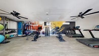 The fitness center at Indulge at Sunset Park, located at 1423 Reno in Las Vegas, NV, is a compact yet fully equipped exercise facility designed to support a well-rounded workout routine. The room features light wood-style flooring, neutral-toned walls, and a textured ceiling with ceiling fans to maintain air circulation throughout the space. A striking, full-wall motivational mural anchors the room, featuring bold fitness-themed typography and dynamic imagery that energizes the environment. A wall-mounted flat-screen TV is positioned above the dumbbell rack for added convenience during workouts. The facility is outfitted with a diverse selection of equipment, including stationary bikes, a treadmill, an elliptical trainer, a multi-station cable machine, a dumbbell rack, medicine balls, stability balls, and kettlebells — providing options for both cardiovascular and strength training. Glass entry doors allow natural light to filter into the space, creating an open and welcoming atmosphere. A bulletin board near the entrance displays posted notices for residents. The thoughtful layout maximizes the available floor area while keeping traffic flow clear and efficient, making this fitness center a practical and motivating amenity for residents.