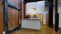 This single-floor loft showcases a striking blend of industrial heritage and modern functionality. The space is immediately defined by its dramatic ceiling height, with towering exposed brick walls that span the full vertical reach of the interior, lending a sense of grandeur and raw authenticity. Dark steel structural beams and columns punctuate the space, serving as bold architectural reminders of the building's historic character.

The open-concept kitchen sits at the heart of the living area, anchored by a peninsula with a dark countertop that provides ample prep and casual dining space. Light wood cabinetry lines the walls, complemented by stainless steel appliances including a refrigerator, microwave, and range. A decorative pendant chandelier hangs above, adding a touch of warmth to the otherwise industrial aesthetic.

Rich hardwood flooring flows throughout the main living area, enhancing the warmth of the space. A compact bathroom with a toilet, sink, and wall-mounted medicine cabinet is tucked neatly off the main corridor, efficiently utilizing the available layout. Additional doorways suggest further rooms or storage beyond the main living zone. Overall, the space delivers a compelling urban loft atmosphere defined by texture, height, and timeless industrial design elements.