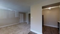 This single-floor space is designed with a practical, open layout that connects the main living area to the kitchen and additional rooms. The living area features wall-to-wall carpeting in a neutral tone, contributing to a warm and understated ambiance. Smooth beige walls are complemented by white baseboards and trim, creating a clean, cohesive look throughout. A textured popcorn ceiling adds a classic residential feel to the interior.

Natural light enters through horizontal blind-covered windows along one wall of the living area, providing brightness while maintaining privacy. The front entry door is centrally positioned, offering straightforward access to the main living space. A doorway adjacent to the entry leads to an additional room or hallway, suggesting a functional division between living and private areas.

Transitioning toward the kitchen, the flooring shifts to dark hardwood-style planks, clearly delineating the two zones. A stainless steel refrigerator is visible within the kitchen area, indicating a well-equipped culinary space. Overall, the layout is efficient and versatile, suited for comfortable everyday living.