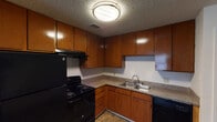 This single-story space is designed for practical, comfortable living. The kitchen is well-equipped, featuring warm medium-toned wood cabinetry that extends to the upper walls, offering generous storage space. Speckled laminate countertops run along the L-shaped layout, complemented by a double stainless steel sink, a black electric range with overhead ventilation, and a black dishwasher. Wood-look flooring and a textured ceiling complete the kitchen's functional character.

The living area is carpeted and bathed in natural light from a window fitted with horizontal blinds. A ceiling fan with a brass finish provides both lighting and air circulation, contributing to the room's relaxed atmosphere. The textured ceiling continues throughout the space, lending a consistent aesthetic.

The bedroom is spacious, carpeted, and includes a window that allows natural light in. It connects to a generously sized walk-in closet outfitted with dual hanging rods, providing ample storage and organization potential.

The bathroom features a wood-toned vanity with a speckled countertop, an oval undermount sink, a wide mirror with overhead lighting, and a separate toilet and tub-shower combination with white tile surround.