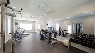 Located at 100 Lake Clair Place in Fayetteville, NC, this fitness center offers a well-appointed workout environment designed to support a range of exercise routines. The single-story space features light-toned walls and recessed lighting complemented by multiple ceiling fans, which help maintain airflow and comfort throughout sessions. Large windows along the perimeter allow natural light to fill the room, creating an open and energizing atmosphere. Full-length mirrors are mounted along one wall, visually expanding the space and providing users with real-time feedback during workouts. The flooring consists of durable, dark rubberized material suited for heavy equipment and high-traffic use. The facility is outfitted with a solid selection of strength-training machines, free weights, a stability ball, and cardiovascular equipment including a treadmill and elliptical-style machines. The layout is thoughtfully organized, providing ample room to move between stations without feeling cramped. Overall, the space presents a clean, functional, and well-maintained environment suitable for a variety of fitness levels and workout styles.