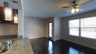 This single-floor space showcases a clean, modern aesthetic defined by its striking polished concrete floors, which reflect light and lend a sleek, industrial character to the interior. The open layout creates a seamless flow between the living and kitchen areas, with a granite countertop visible along one edge, hinting at a well-appointed kitchen space. Neutral gray walls keep the atmosphere bright and versatile, providing a blank canvas adaptable to a variety of uses. Track lighting along the ceiling offers focused, adjustable illumination, while a ceiling fan with a built-in light fixture adds both comfort and additional lighting to the main living area. Natural light filters in through multiple windows, including one that frames an outdoor view of surrounding greenery, enhancing the airy feel of the space. A door with a glass panel provides direct access to the exterior, further connecting the interior to the outdoors. The combination of modern finishes, functional lighting, and an open, flowing layout makes this a practical and stylish single-floor space suited for a range of purposes.