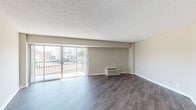 This well-proportioned two-bedroom, one-bathroom apartment delivers a practical and comfortable single-floor layout. The spacious living room is a standout feature, showcasing dark hardwood-style flooring, a textured ceiling, and a wall-unit HVAC system. Large sliding glass doors flood the room with natural light and open directly onto a private balcony that overlooks the surrounding urban skyline — an ideal spot for taking in city views. A dedicated dining or secondary living area is accessible via a connecting hallway, also finished with the same rich hardwood-style floors, lending visual continuity throughout. The kitchen is efficiently laid out with white cabinetry, granite-style countertops, a full suite of white appliances including a refrigerator and electric range, and dark hardwood-style flooring. Both bedrooms are generously sized and feature plush wall-to-wall carpeting, neutral tones, and ample closet space with bifold doors. The shared full bathroom includes a granite-style vanity countertop, a large mirror, and a tub-shower combination. Throughout the unit, neutral gray walls and clean white trim create a calm, move-in-ready aesthetic suited for a wide range of interior styles.