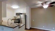 This single-story space offers a practical and well-appointed layout designed for comfortable everyday living. The kitchen is a central highlight, equipped with stainless steel appliances including a gas range, over-the-range microwave, and a full-size refrigerator. White raised-panel cabinetry extends to the ceiling, maximizing storage capacity while lending a clean, classic aesthetic. Dark countertops provide a subtle contrast to the bright cabinetry, adding visual depth to the kitchen's design. A flush-mount ceiling light provides focused illumination over the kitchen workspace.

The kitchen flows openly into an adjoining room, likely a dining or living area, which features warm-toned beige walls and wood-style flooring that adds warmth underfoot. A ceiling fan is visible in the adjacent space, contributing to air circulation and comfort. The textured ceiling is consistent throughout, adding a traditional residential character. The overall color palette of neutral beige tones and white trim creates a cohesive, inviting atmosphere. The open sightlines between the kitchen and adjoining area enhance the sense of connectivity and spaciousness throughout the home.