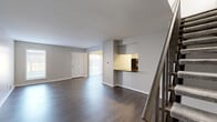 This two-floor residence offers a well-organized layout with distinct living spaces across both levels. The main floor features a spacious living area with dark hardwood flooring, neutral gray walls, and clean white trim throughout. A staircase with a metal railing connects the two floors, while a pass-through window opens into the kitchen, creating a connected feel between the living and cooking areas. The kitchen is well-appointed with white shaker-style cabinetry, dark countertops, stainless steel appliances including a refrigerator, electric range, and dishwasher, complemented by light hardwood-style flooring. A front entry area on the upper level provides a welcoming space with hardwood flooring, a window for natural light, and direct access to the front door. The living area also features a sliding glass door fitted with vertical blinds, opening to an outdoor view and bringing in ample natural light. Bedrooms are finished with plush carpeting and generous closet space, including a walk-in closet with built-in shelving. The bathroom includes a full-size soaking tub with a surround featuring integrated shelving. Overall, the space presents a clean, modern aesthetic with practical finishes throughout.