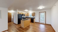 This efficient single-floor unit offers a well-organized layout comprising a living room, kitchen, bedroom, and bathroom. The kitchen serves as a functional centerpiece, featuring light maple cabinetry, dark granite countertops, a full suite of black appliances including a refrigerator, range, microwave, and dishwasher, and a central island that provides additional prep and storage space. Hardwood-style flooring runs throughout the kitchen and living areas, adding warmth to the space. A side door adjacent to the kitchen sink offers convenient exterior access with natural light from multiple windows. The living room is open and straightforward, with hardwood-style flooring continuing from the kitchen and a clean, neutral palette. The bedroom is carpeted and comfortably scaled, featuring a ceiling fan with light fixture, a window with outdoor views, and a closet with direct access to the hallway. The bathroom is finished with tile flooring, a wood-toned vanity with a white countertop, a large mirror, and brushed nickel fixtures and hardware. Textured ceilings and warm wood door trim are consistent throughout the unit, lending a cohesive and practical character to the overall space.