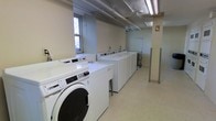 This shared laundry facility is designed with practicality and efficiency in mind. The room features a row of commercial-grade front-loading washing machines and top-loading machines, numbered for easy identification and organized along the walls to maximize available floor space. The light-tiled flooring and neutral-toned walls contribute to a clean, utilitarian aesthetic throughout the space. Overhead fluorescent lighting ensures the room is well-lit, supplemented by natural light filtering in through windows along one wall. Exposed ceiling infrastructure, including pipes and conduits, reflects the functional nature of the facility. A utility cart is also present, providing additional convenience for users transporting laundry. The layout is open and spacious, allowing comfortable movement between machines. Overall, this laundry room is well-equipped to serve the needs of a multi-unit or communal building, offering a straightforward and accessible environment for residents or occupants.