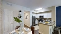 This single-floor residence offers a thoughtfully designed open-concept layout that seamlessly connects the kitchen and dining areas. The kitchen is well-appointed with stainless steel appliances, including a refrigerator, gas range, and built-in microwave, all complemented by crisp white cabinetry and a light-toned countertop island that doubles as a functional workspace and casual gathering spot. A stainless steel sink with a modern faucet adds to the kitchen's clean, contemporary aesthetic. The dining area is positioned adjacent to the kitchen, creating a natural flow ideal for everyday living and entertaining. The space is finished with rich wood-look flooring that adds warmth and contrast against the bright white walls. Recessed and overhead lighting ensures the interior remains well-lit throughout. A hallway leads further into the residence, lined with decorative built-in shelving displaying potted plants that introduce a touch of greenery and life to the space. Framed wall art along the corridor adds a curated, welcoming ambiance. The overall design combines modern finishes with a neutral color palette, creating a clean, bright, and inviting living environment.