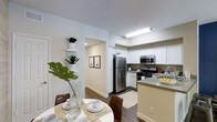 This single-floor residence offers a thoughtfully designed open-concept layout that seamlessly connects the kitchen and dining areas. The kitchen is well-appointed with stainless steel appliances, including a refrigerator, gas range, and built-in microwave, all complemented by crisp white cabinetry and a light-toned countertop island that doubles as a functional workspace and casual gathering spot. A stainless steel sink with a modern faucet adds to the kitchen's clean, contemporary aesthetic. The dining area is positioned adjacent to the kitchen, creating a natural flow ideal for everyday living and entertaining. The space is finished with rich wood-look flooring that adds warmth and contrast against the bright white walls. Recessed and overhead lighting ensures the interior remains well-lit throughout. A hallway leads further into the residence, lined with decorative built-in shelving displaying potted plants that introduce a touch of greenery and life to the space. Framed wall art along the corridor adds a curated, welcoming ambiance. The overall design combines modern finishes with a neutral color palette, creating a clean, bright, and inviting living environment.