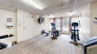 Located at 262 Rose St in Barrie, Ontario, this dedicated exercise room offers a practical and well-equipped space for residents to maintain an active lifestyle. The room features a drop tile ceiling with recessed fluorescent lighting that keeps the area well-lit throughout. Patterned, low-pile carpeting covers the floor, providing a functional surface suited for cardio-focused workouts.

The space is outfitted with a variety of Life Fitness cardio equipment, including treadmills, elliptical cross-trainers, a stationary upright bicycle, a rowing machine, and a stair climber, offering a comprehensive range of cardiovascular exercise options. A wall-mounted television is positioned near the exit door, allowing users to stay entertained during workouts.

Natural light filters into the room through multiple windows fitted with vertical blinds, offering views of the surrounding outdoor greenery and creating a pleasant, airy atmosphere. Baseboard heating units are positioned along the walls, ensuring comfort during cooler months. Posted hours-of-operation signage and decorative framed artwork add a welcoming touch to the otherwise utilitarian space. The room's compact yet efficient layout makes thoughtful use of the available floor area to accommodate multiple users simultaneously.