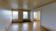 This single-floor space showcases a blend of classic architectural character and functional design. The main room is spacious and airy, highlighted by warm hardwood floors that run throughout, reflecting natural light and adding a sense of warmth and continuity. Tall, multi-pane windows line the exterior wall, flooding the room with abundant daylight while offering views of the surrounding outdoor scenery.

Architectural detailing is evident throughout, including crown molding that frames the ceilings and substantial baseboards that complement the period-inspired aesthetic. The ceilings are generously proportioned, contributing to an open and comfortable feel. Cast-iron radiators beneath the windows speak to the building's heritage and provide a charming, functional element.

An open doorway connects the main room to an adjoining space, which shares the same hardwood flooring, white walls, and natural light from its own window. The consistent palette of neutral tones and natural wood creates a cohesive, clean ambiance throughout. Overall, the space presents a well-preserved, classic interior with timeless architectural features suitable for a variety of uses.