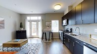 This well-designed single-floor residence seamlessly blends the kitchen, dining, and living areas into a cohesive open-concept layout. The kitchen is a standout feature, boasting rich dark cabinetry that contrasts beautifully with light granite countertops. Stainless steel appliances, including a range and microwave, provide a polished, contemporary finish. Ample cabinet storage ensures both functionality and style throughout the cooking space. Adjacent to the kitchen, a sleek bar-height dining table with industrial-style stools creates an inviting casual dining nook. The living area extends naturally from the dining space, anchored by wood-style flooring that runs throughout and adds warmth to the neutral-toned walls. A bold geometric area rug defines the living zone and adds a decorative touch. Natural light floods the interior through multiple windows and a set of glass French doors that open to an outdoor area, creating a bright and airy ambiance. The doors are dressed with flowing white curtains that soften the space. The overall aesthetic is modern and clean, combining dark and light tones with functional furnishings to deliver a comfortable and stylish living environment.