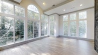 This single-floor space makes an immediate impression with its airy, open layout and thoughtfully designed architectural details. The centerpiece of the main living area is a striking arched window that crowns a wall of floor-to-ceiling glass panels, flooding the interior with abundant natural light and offering sweeping views of the surrounding mature trees and greenery. The vaulted ceiling adds a sense of grandeur and vertical openness, enhancing the spacious feel of the room. Recessed lighting complements the natural illumination, ensuring the space remains bright at any hour. Elegant crown molding and a tray ceiling detail add a refined architectural touch, lending the space a polished, upscale character. The light-toned hardwood flooring runs throughout, contributing warmth and a clean, cohesive aesthetic that ties the interior together seamlessly. The neutral white and gray palette of the walls and trim creates a versatile backdrop, well-suited for a variety of interior design styles. Corner window arrangements on multiple walls maximize the connection between the interior and the lush natural surroundings, making the outdoors feel like a natural extension of the living space. Overall, the space exudes a calm, luminous ambiance with a strong emphasis on natural light, open volume, and refined finishes.