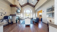 This single-floor community clubhouse is a thoughtfully designed gathering space that immediately impresses with its dramatic vaulted ceiling, accented by exposed dark wood beams that add warmth and architectural character. Rich hardwood-style flooring flows throughout, complementing the open and airy atmosphere created by the soaring ceiling heights.

The main lounge area serves as the heart of the space, featuring a striking floor-to-ceiling stacked stone fireplace flanked by a wall-mounted flat-screen television. Comfortable seating arrangements anchored by a bold chevron-patterned area rug invite relaxation and socializing. A grand arched window at the far end floods the room with natural light, offering outdoor views and creating a bright, welcoming ambiance.

The entry foyer is equally impressive, featuring a graceful arched doorway, built-in display ledges, and a central accent table with a decorative floral arrangement — setting an elegant tone upon arrival.

Adjacent to the lounge, a well-appointed business center provides workstations equipped with computers and a wall-mounted display screen, ideal for residents needing a productive workspace. A dedicated leasing or service counter area features dark cabinetry, a stone accent wall, and display shelving, providing a polished and functional point of contact. Together, these areas create a cohesive, modern clubhouse that balances comfort, style, and practicality.