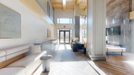 This elegantly designed single-floor space offers a generous footprint that seamlessly connects a refined living room and a well-appointed dining room. The most striking architectural feature is the dramatic double-height ceiling in the living area, accented by exposed natural wood beams and clerestory windows that flood the interior with abundant natural light. Large floor-to-ceiling glass doors and windows provide views of the surrounding exterior, enhancing the open, airy atmosphere throughout.

The living room showcases herringbone-patterned hardwood flooring, substantial structural columns with detailed millwork, and upper-level paneled cabinetry in a muted sage-gray tone. A bold floor-to-ceiling marble tile accent wall anchors the space alongside a wall-mounted television, while the seating area is appointed with contemporary furnishings.

The dining room continues the herringbone flooring and features a slatted gray ceiling detail, a sculptural multi-globe brass chandelier, and a large oval dining table surrounded by mixed wood-and-leather chairs. A built-in wet bar or service counter with patterned tile backsplash runs along one wall, adding functional elegance. Large windows with sheer drapery allow natural light to pour in, completing this refined and welcoming communal environment.