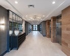 The lobby at 940 Stuyvesant Ave in Union, New Jersey, presents a thoughtfully designed first-floor common area that blends modern aesthetics with functional amenities. The grand entrance corridor features large-format light stone tile flooring that extends throughout, creating a seamless and refined foundation underfoot. A statement brass sputnik chandelier anchors the ceiling above the main hallway, complemented by recessed lighting and elegant wall sconces that maintain a warm, welcoming ambiance throughout the day.

Wood-clad accent walls add natural warmth and texture, contrasting beautifully against the cool grey and dark charcoal wall finishes. A pair of stainless steel elevators are framed by striking stone-tiled surrounds, offering residents convenient vertical access. Along one side of the corridor, a sleek black console table and mirrored glass partition add a decorative focal point, while floor-to-ceiling glass entry doors invite natural light deep into the space and offer views of the surrounding streetscape.

The mailroom area is outfitted with golden multi-unit mailboxes mounted on a grey accent wall, paired with a wood-panel reception counter beneath the building's branded logo medallion. A cozy lounge nook with a round bistro table, mixed seating, and a pendant light sits beside expansive windows, providing residents a comfortable spot to relax while overlooking the exterior.