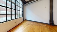 Unit 316 at 101 E Sixth St offers a loft-inspired aesthetic in the heart of downtown Winston-Salem. Original hardwood floors and exposed ductwork carry throughout the open layout, reflecting the building's industrial character. Floor-to-ceiling black-framed windows flood the living and bedroom areas with natural light and offer views of the surrounding brick architecture. The galley kitchen is well-appointed with dark espresso cabinetry, granite countertops, stainless steel appliances, and a double sink. The bedroom includes dual closets with built-in wire shelving for ample storage. The full bathroom features large-format tile flooring, a soaking tub with overhead shower, a dark wood vanity, and granite countertops.